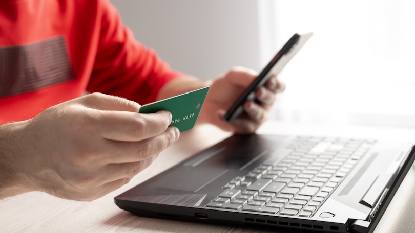 Eine Person sitzt an einem Schreibtisch vor einem geöffneten Laptop und hält in der rechten Hand eine Kreditkarte sowie in der linken Hand ein Smartphone, um sich auf einen Online-Bezahlvorgang vorzubereiten. Eine Person sitzt an einem Schreibtisch vor einem geöffneten Laptop und hält in der rechten Hand eine Kreditkarte sowie in der linken Hand ein Smartphone, um sich auf einen Online-Bezahlvorgang vorzubereiten.