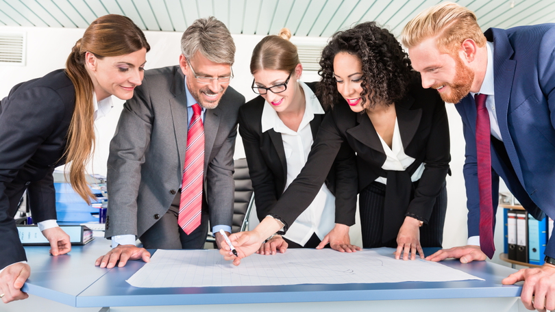 Eine Gruppe von Personen steht gemeinsam an einem Konferenztisch und arbeitet an einem Dokument Eine Gruppe von Personen steht gemeinsam an einem Konferenztisch und arbeitet an einem Dokument