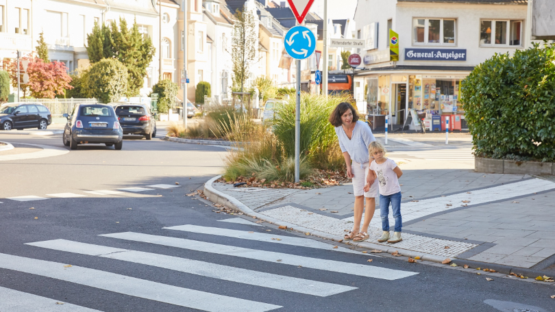 Eine Frau und ein Kind stehen an einem Zebrastreifen und prüfen mit einem Blick nach links, ob die Straße sicher überquert werden kann Eine Frau und ein Kind stehen an einem Zebrastreifen und prüfen mit einem Blick nach links, ob die Straße sicher überquert werden kann